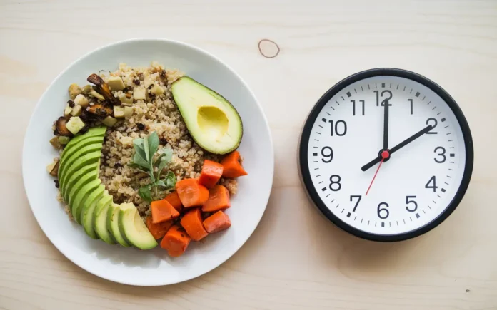 A vibrant plate of vegan food, including quinoa, avocado, and colorful vegetables, paired with a clock symbolizing intermittent fasting.